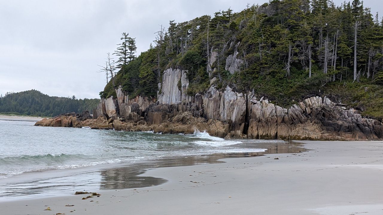 Dramatic rocky cliffs topped with evergreens meeting the sandy beach at Pruth Bay