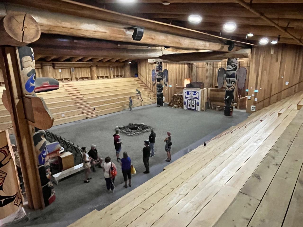 Interior of the big house in Bella Bella with massive cedar beams, totem poles, and visitors gathered around the central fire pit