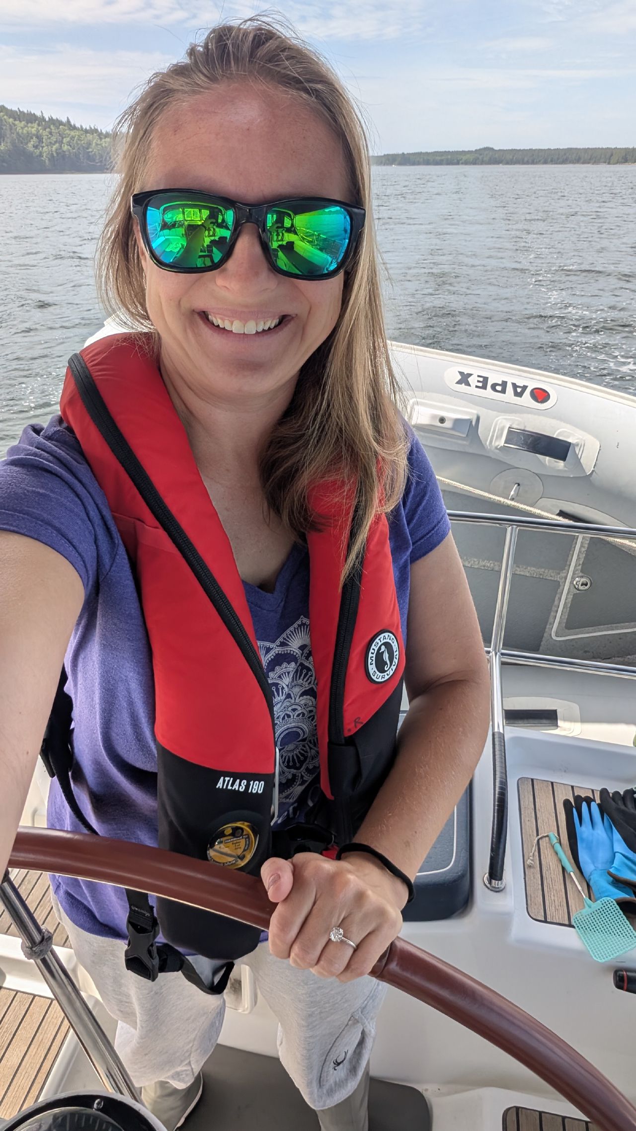 Rachel smiling at the helm in sunglasses and life jacket on a beautiful sunny passage day