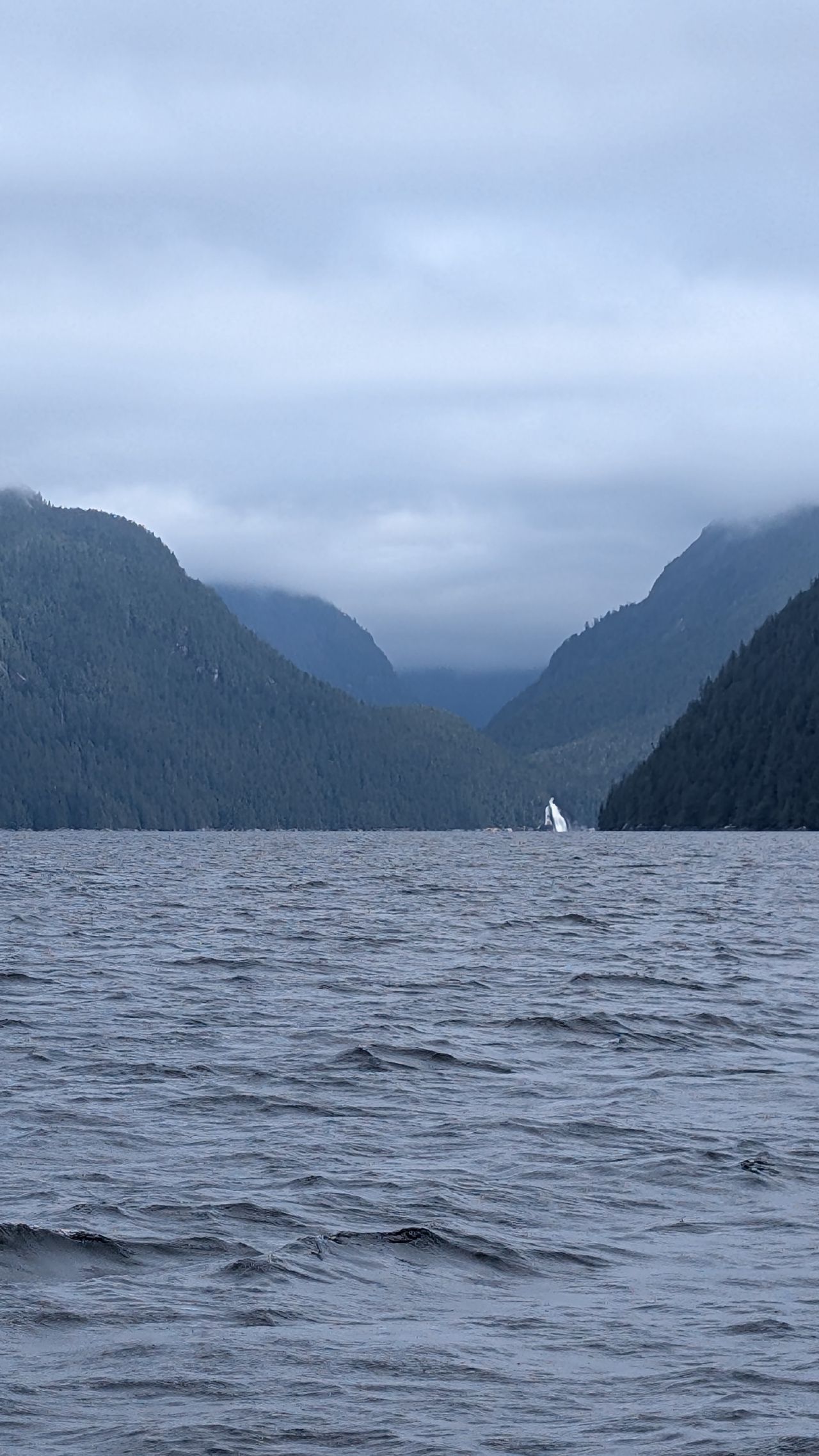 Misty mountain valley with a distant waterfall framed by forested peaks and choppy water