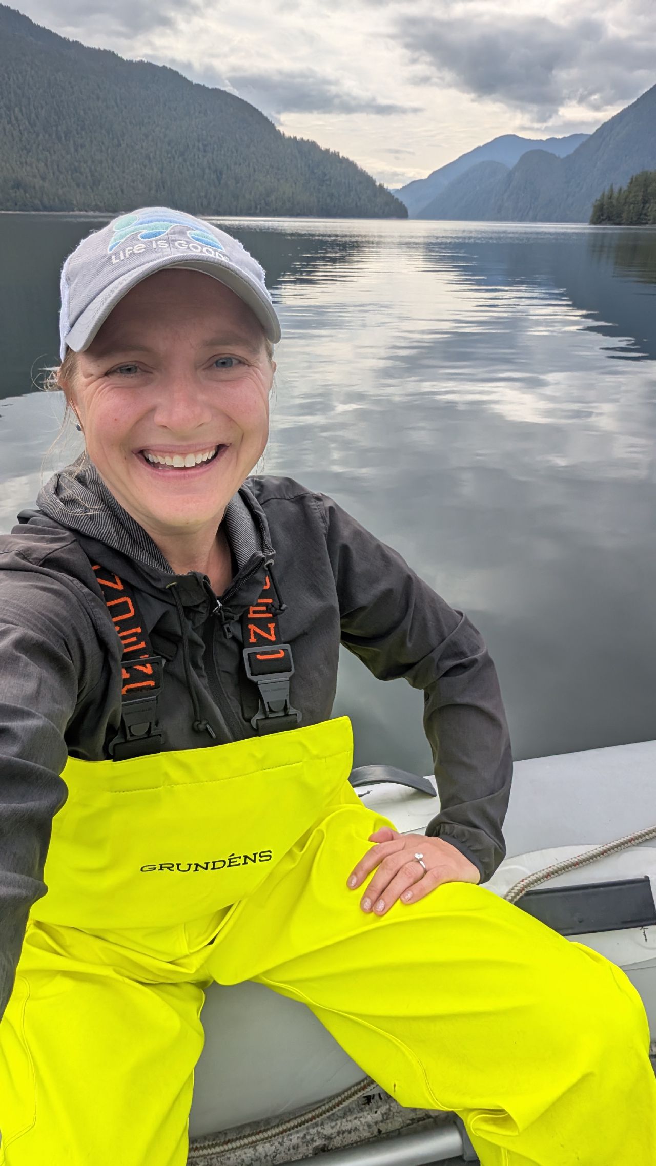 Rachel selfie in yellow Grundens rain gear in the dinghy with glassy water and mountains behind