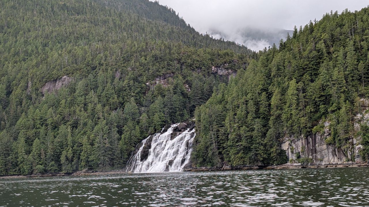 Wide waterfall pouring down the forested mountainside directly into the ocean at Poison Cove