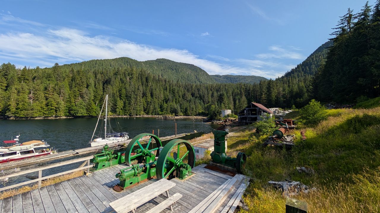 Old green cannery machinery on the dock at Butedale with sailboats and mountains in the background