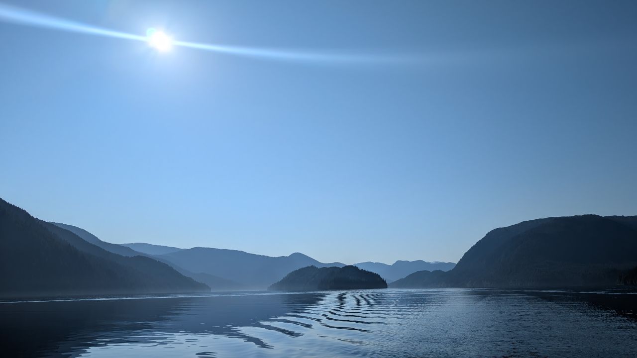 Hazy sun over layered mountain silhouettes and glassy water en route to Hartley Bay