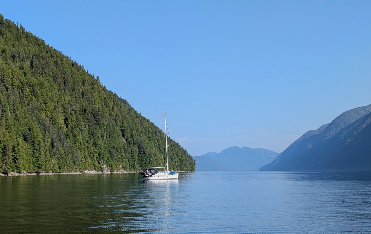 A sailboat motoring through a calm channel flanked by forested mountains on the way to Hartley Bay