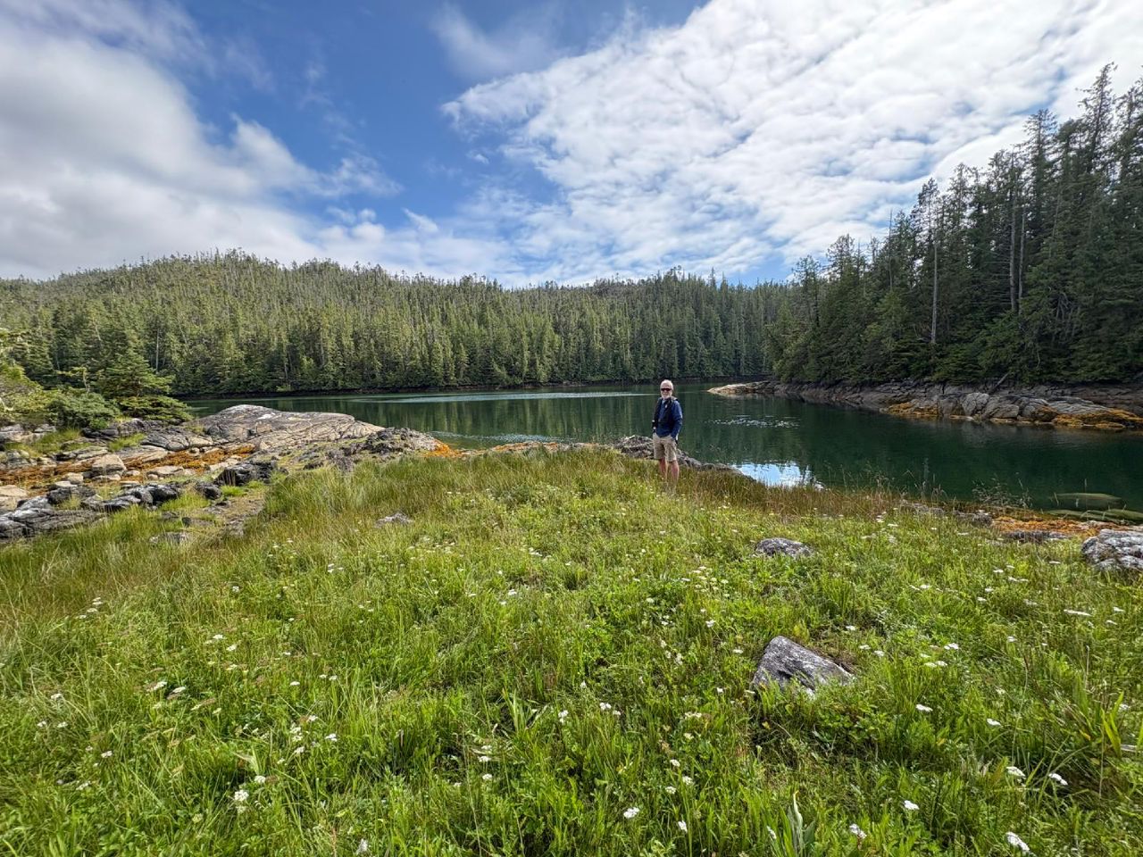 Brian standing on a green meadow at the edge of a tranquil cove surrounded by evergreen forest