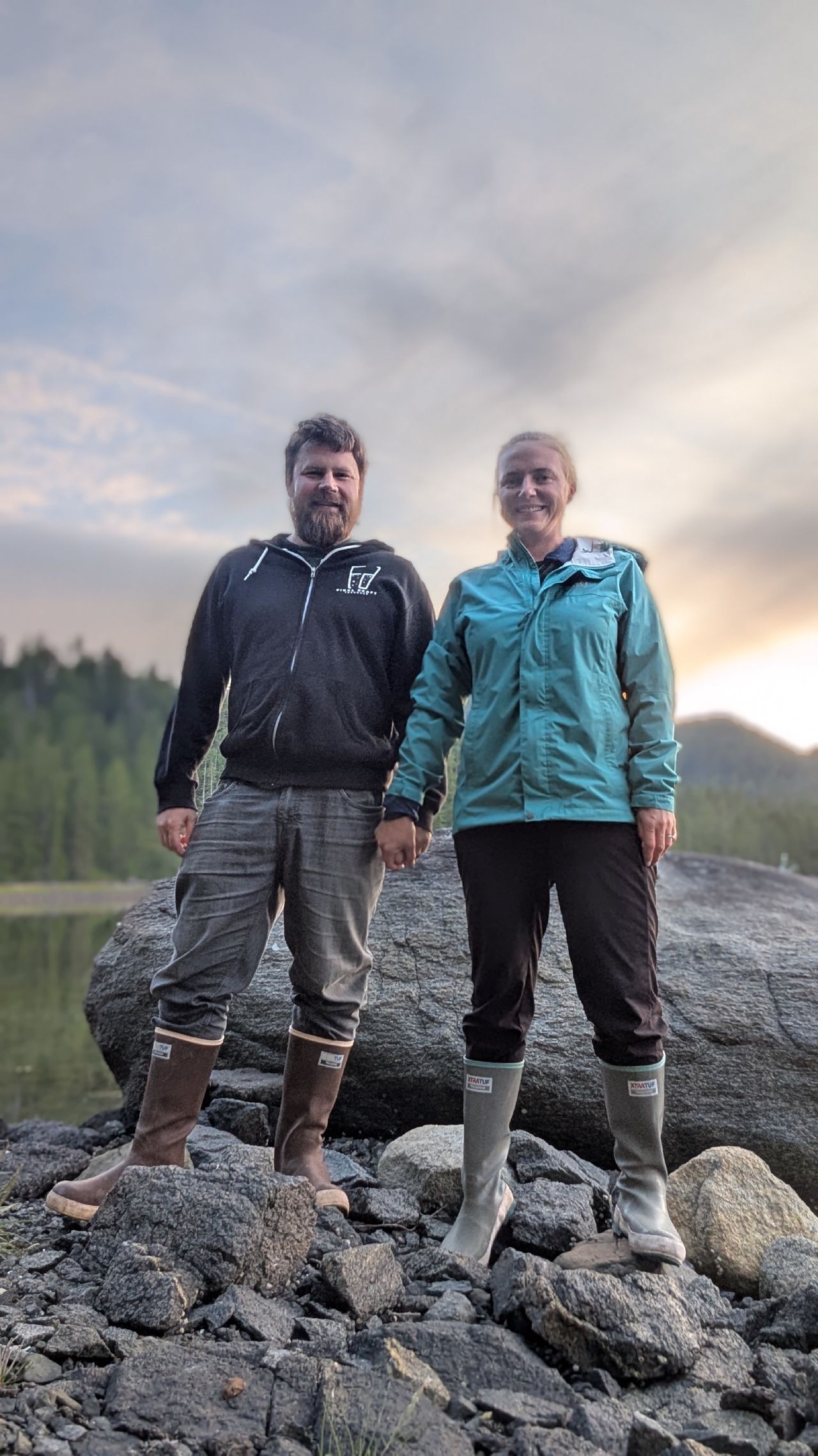 Brian and Rachel standing together on rocks in rubber boots with a sunset sky behind them