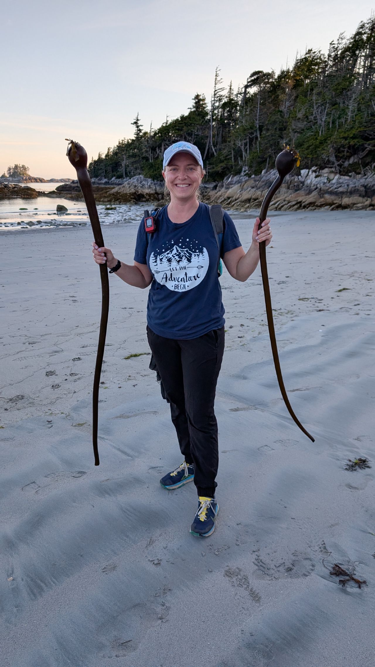 Rachel smiling and holding two large pieces of bull kelp on the beach at Pruth Bay