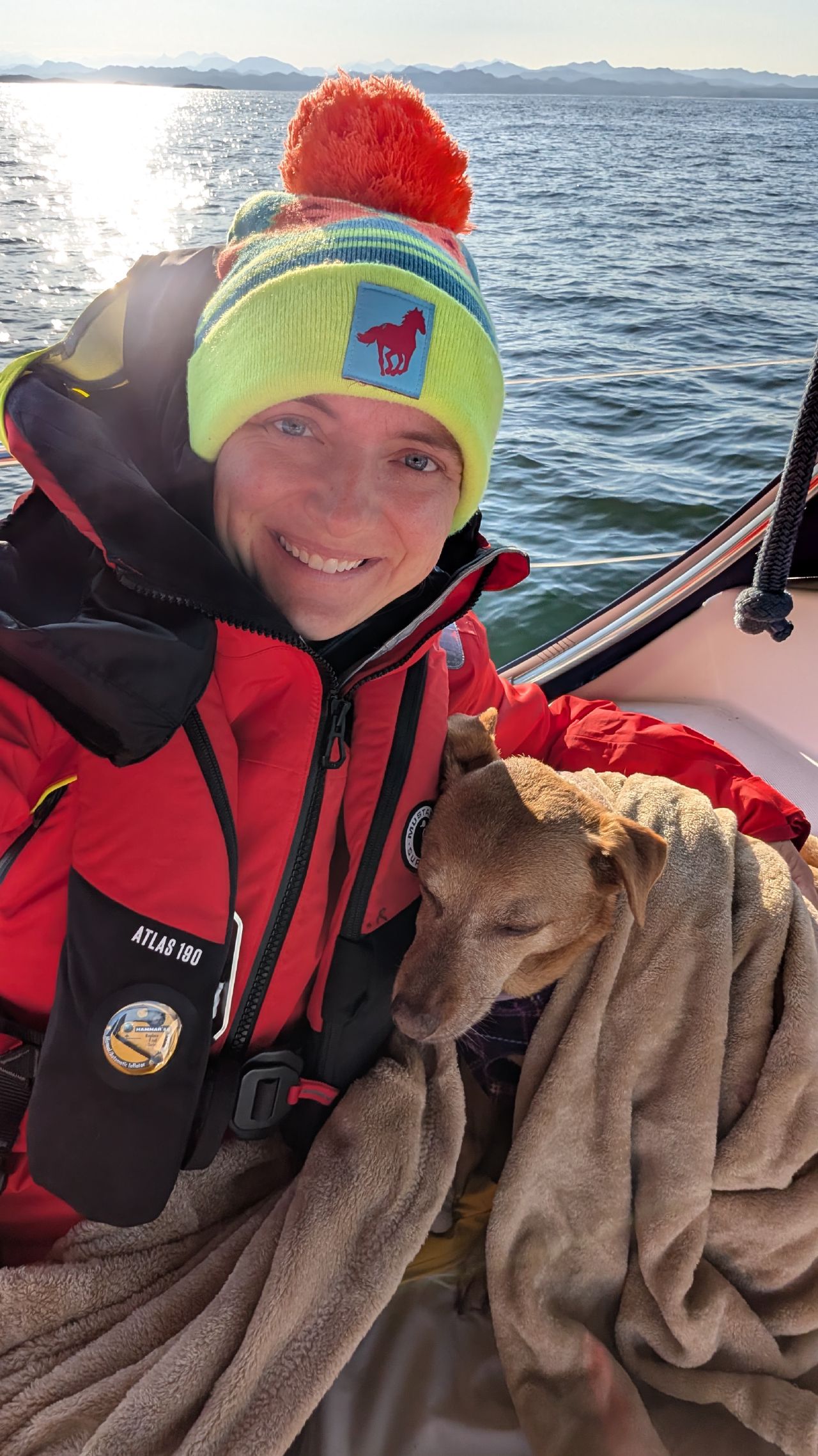 Rachel smiling in her red foul weather gear and colorful beanie with Maui wrapped in a blanket in the cockpit