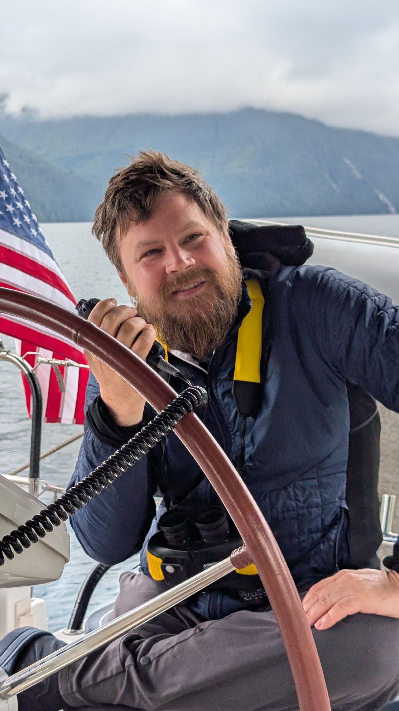 Brian smiling at the helm of SV Yahara with the American flag and cloud-covered mountains behind