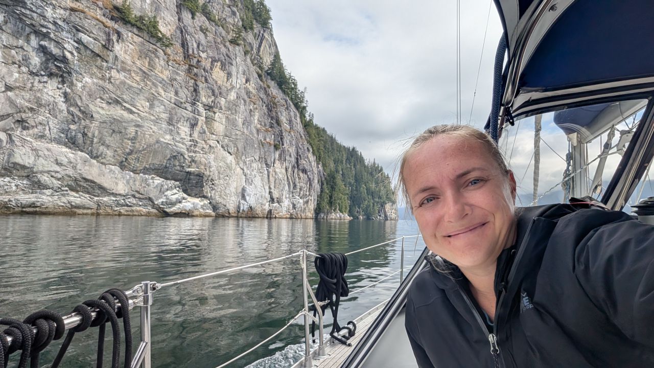 Rachel taking a selfie on the bow of SV Yahara passing a dramatic granite cliff face