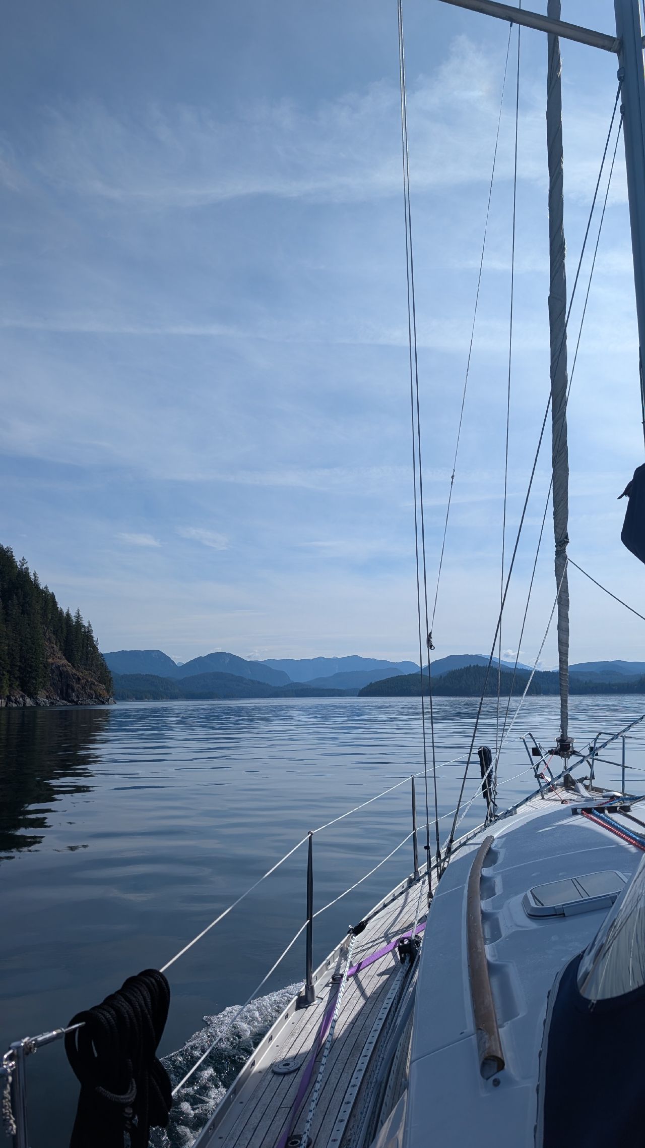 View along the teak deck of SV Yahara motoring through calm waters with BC mountains in the distance
