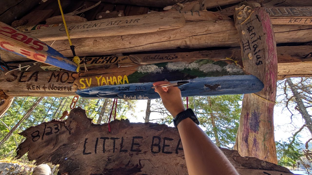 Hand pointing to the painted SV Yahara driftwood art piece with 2025 newly added in the driftwood cabin