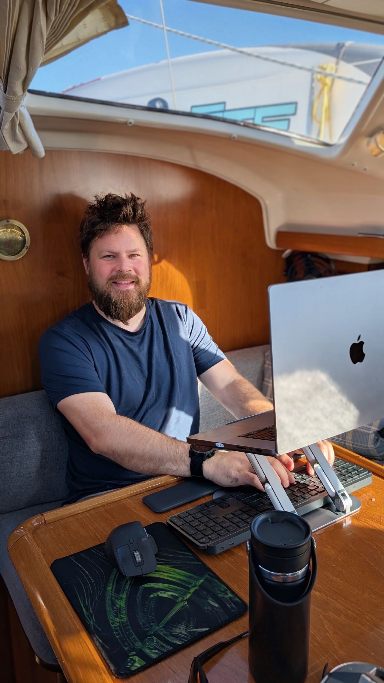 Brian smiling at his remote work setup with laptop and keyboard at the saloon table after anchoring at Rebecca Spit