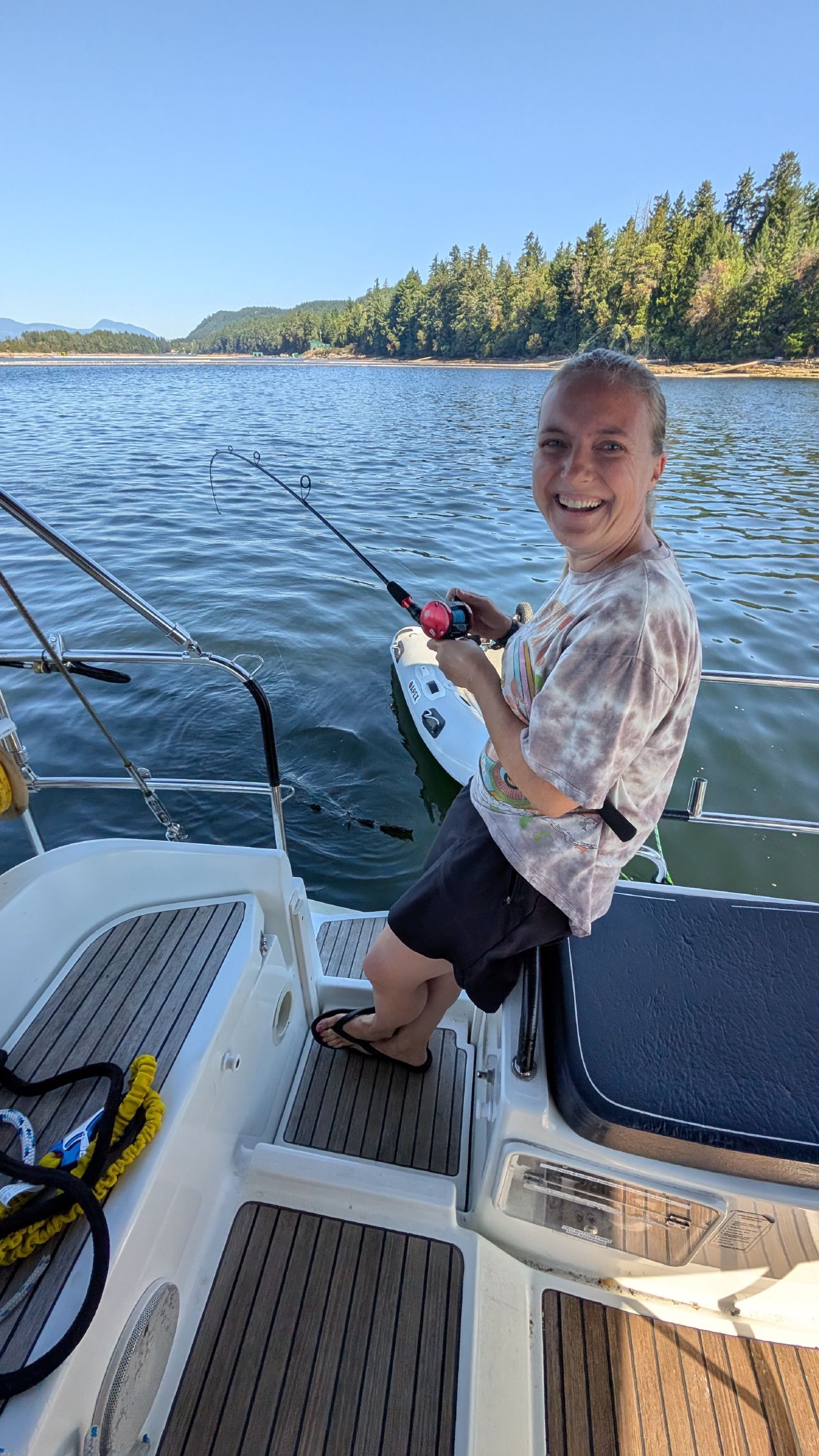 Rachel smiling while holding a fishing rod off the stern of SV Yahara in a calm Ladysmith anchorage