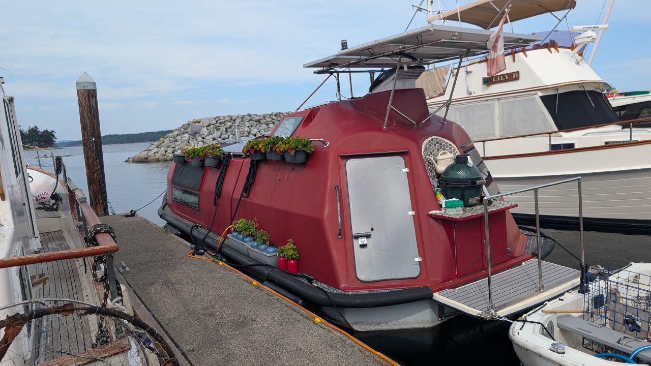 Red converted ferry lifeboat turned into a liveaboard home with solar panels and potted plants at Sidney marina