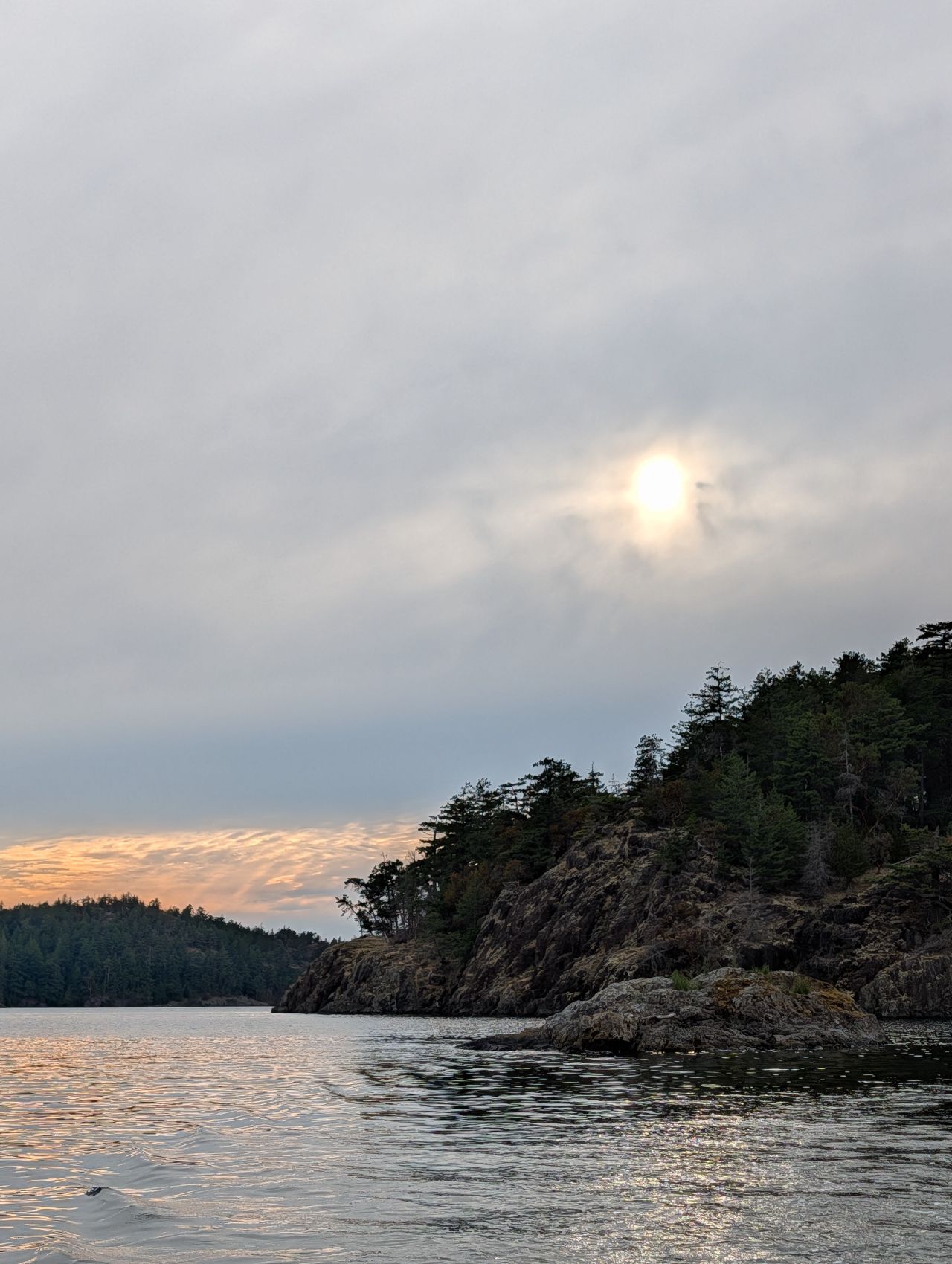 Hazy sun setting behind a rocky, tree-lined point